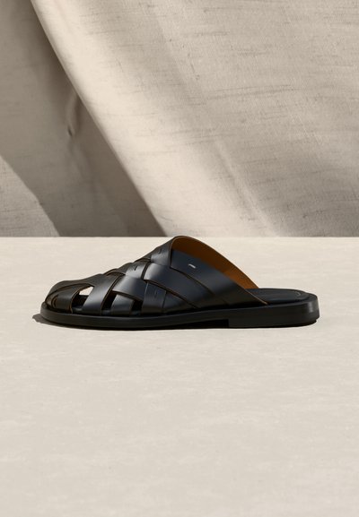 Sandale noire en cuir à enfiler avec un design tissé et une semelle plate, présentée sur une surface neutre avec un fond en tissu beige.