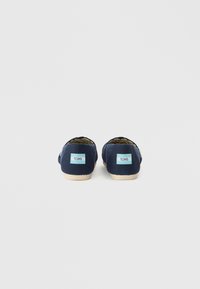 Granatowe płócienne buty wsuwane z jasnokremowymi podeszwami. Akcent z logo TOMS w jasnoniebieskim kolorze na tyle. Prosty, zaokrąglony design. Teksturowany materiał.