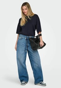 Haut bleu marine oversize à manches courtes, jean large en denim bleu, et sac à main en cuir noir. Les baskets présentent des détails bleus et blancs.