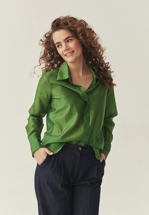 Žena s kovrčavom kosom koja nosi jarko zelenu bluzu i tamnoplave hlače, nasmiješena i blago okrenuta u stranu, ruke u džepovima.