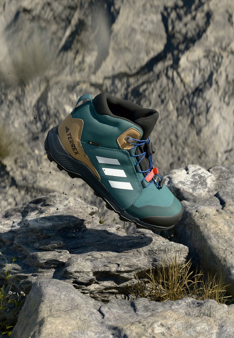 Botas de hiking turquesa com colarinho preto, detalhes castanhos, riscas brancas e atacadores azuis. Elevadas acima de um terreno rochoso com relva verde abaixo.