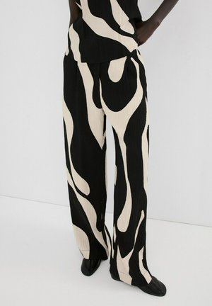 Nohavice s širokými nohavicami s čiernymi a béžovými abstraktnými vírivými vzormi, nosené s topom v rovnakom prevedení a čiernymi topánkami.