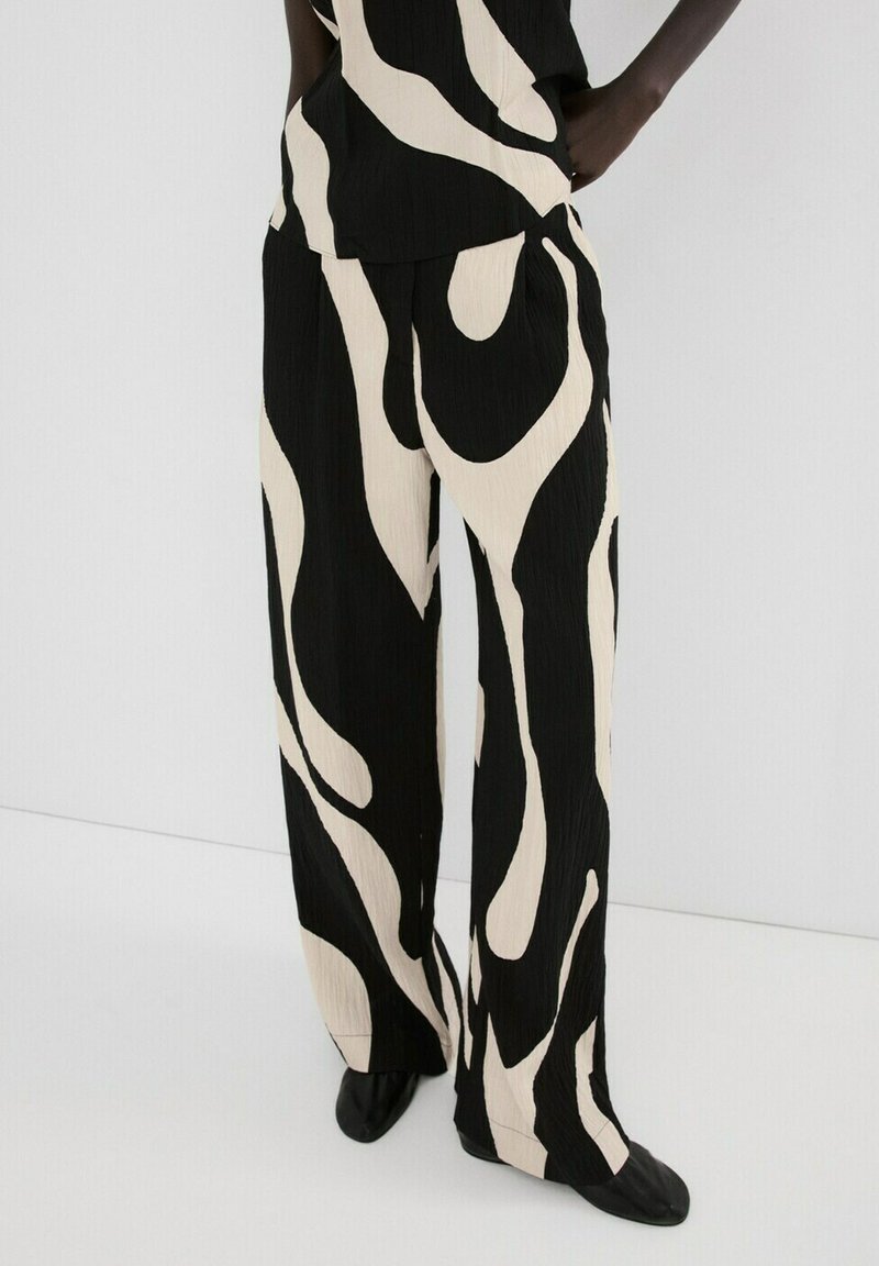 Kalhoty s širokými nohavicemi s černými a béžovými abstraktními spirálovými vzory, nošené s odpovídajícím topem a černými botami.