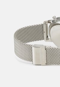 Bracelet de montre en mailles argentées avec un motif texturé, dotée d'un fermoir en métal marqué "FOSSIL" et d'un design épuré et minimaliste.