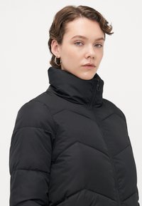 Giovane con capelli ricci corti che indossa orecchini a cerchio argento e un giubbotto imbottito nero con colletto alto zip contro uno sfondo bianco.