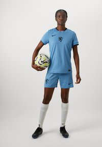 Ljusblå fotbollsdräkt med svart lejonemblem, svart Nike-logga, matchande shorts, vita strumpor och svarta skor. Spelaren håller i en mönstrad fotboll.