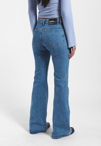 High-waisted flared jeans in medium blue denim, featuring a rough texture, five pockets, and a black brand label at the back.