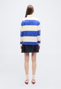 Femme debout, vue de dos, portant une chemise à manches longues rayée bleu et blanc, une jupe foncée, des chaussettes beiges et des sandales à talons rouges.