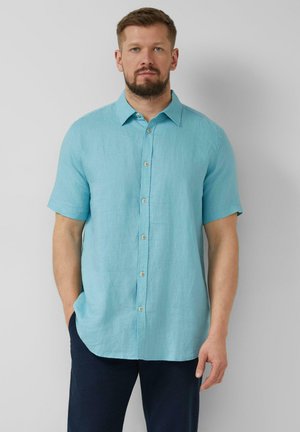 Man draagt een lichtblauw overhemd met korte mouwen en knopen en donkere broek, staat met één hand in de zak tegen een effen achtergrond.