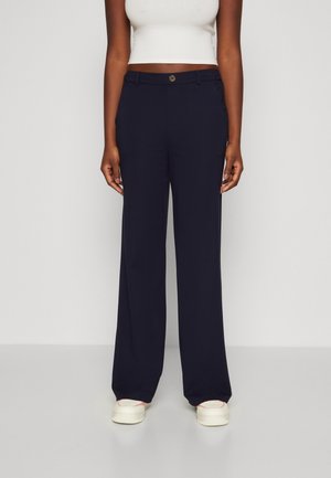 Osoba ubrana w granatowe proste spodnie ze ściągaczem, biały krótki top i białe tenisówki stoi na tle jasnego, jednolitego tła.