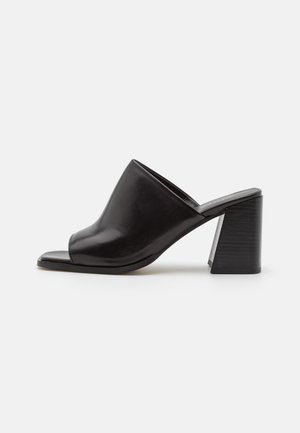 Mules ouvertes en cuir noir avec un dessus incurvé, un talon bloc solide et une texture lisse, présentant un design minimaliste.