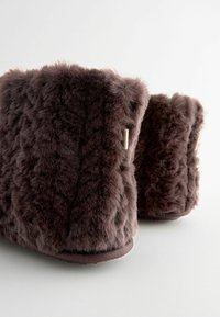 Faux-Fur Hausschuhe in Dunkelbraun, mit einer strukturierten Oberfläche, runder Form und dezenten Seitenakzenten. Weiches und gemütliches Design für den Innenbereich.