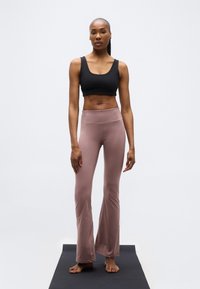 Γυναίκα που στέκεται ξυπόλητη σε μαύρο χαλάκι γιόγκα, φοράει μαύρο αθλητικό κράνος και ροζ-μοβ παντελόνι γιόγκα, μπροστά σε απλό λευκό φόντο.