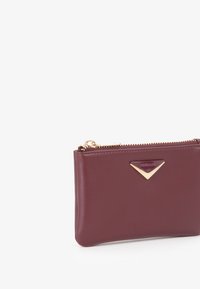 Portafoglio in pelle bordeaux con un accento a triangolo in tono oro, chiusura a zip e texture liscia. Forma compatta e rettangolare per una facile portabilità.