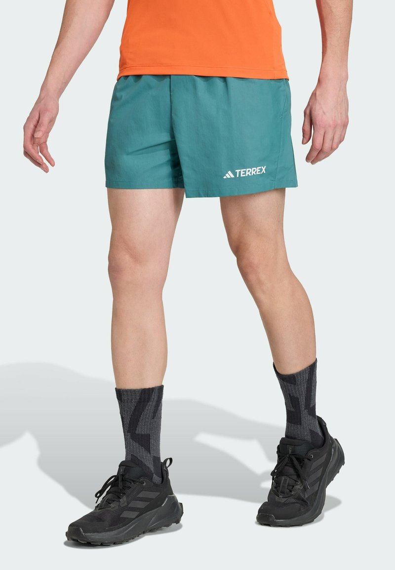 Turquoise sportshorts met een elastische tailleband en een witte "TERREX" logo. Geko paired met zwarte sneakers en gestreepte donkergrijze sokken.