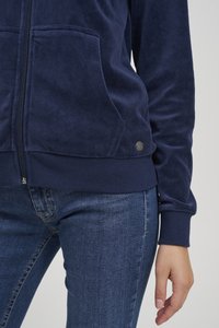 Tmavomodrá mikina na zips z mäkkého materiálu s dvoma prednými vreckami a rebrovanými manžetami. Má malú kovovú nášivku v dolnej ľavej časti.