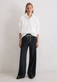 Biała koszula zapinana na guziki z długimi rękawami, w połączeniu z czarnymi spodniami o szerokiej nogawce z elastycznym paskiem i białymi paskami po bokach. Czarne buty.