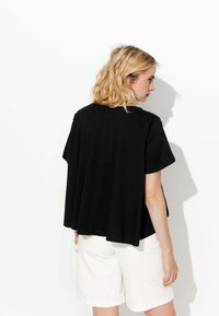 Haut noir à manches courtes en tissu doux avec une coupe ample et fluide. Associé à un short de couleur claire, présenté de dos sur un fond blanc.