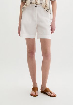 Persoon draagt witte hoog getailleerde op maat gemaakte shorts met knoopsluiting en bruine platte sandalen, staand tegen een effen witte achtergrond.