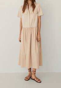 Abito beige con bottoni, maniche corte, due tasche sul petto e vita arricciata, abbinato a sandali marroni con lacci.