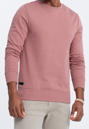 Man wearing a dusty rose long-sleeve crewneck sweatshirt and light beige pants, with a bracelet on his right wrist.