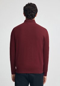 Burgundy stickad tröja med hög krage, långa ärmar och ribbade muddar och fåll. Slät textur, figurnära silhuett.