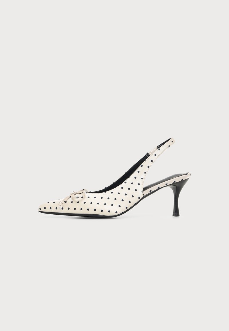 Chaussure à talon haut en cuir blanc à bride arrière avec des pois noirs et un petit nœud décoratif sur l’orteil pointu, sur fond uni.