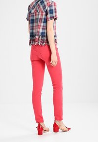 Camisa de cuadros de manga corta con dobladillo con flecos, combinada con jeans ajustados rosas y sandalias rojas de tacón. El conjunto presenta colores vibrantes y patrones distintos.