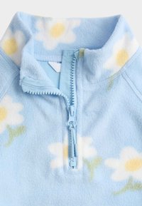 Pile con mezza cerniera in azzurro chiaro con motivi di margherite bianche. Presenta una cerniera frontale con linguetta abbinata e un tessuto morbido e testurizzato.