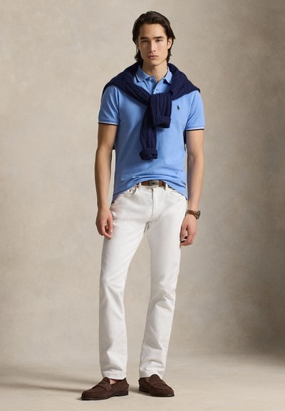 Ανοιχτό μπλε πόλο πουκάμισο, λευκό παντελόνι, μπλε πουλόβερ δεμένο γύρω από το λαιμό, καφέ μοκασίνια και ρολόι χειρός. Το φόντο είναι ουδέτερο.