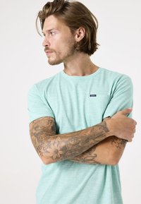 Camiseta de color verde menta de manga corta hecha de una tela suave, con un pequeño bolsillo en el pecho y una textura sutil. El modelo muestra brazos tatuados.