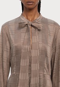 Smeđa bluzica s uzorkom koja ima ovratnik s mašnom, s kombinacijom kariranih i uzoraka houndstooth, prikazujući glatku teksturu tkanine.