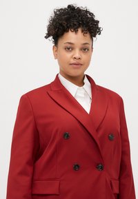 Raudona dvigubos sagos švarkas, pagamintas iš lygaus audinio, su juodomis sagomis ir įpjovimu, derinamas su balta marškinių apykakle po juo.