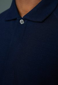 Donkerblauwe polo shirt van gestructureerde stof, met een kraag en een enkele zilveren knop bij de sluiting. Close-up zicht.