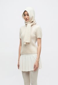 Pulover crem cu mâneci scurte, tricotat, cu fular și glugă asortate; textură netedă, croială lejeră, purtat cu o fustă albă evazată și ciorapi subțiri deschiși la culoare.