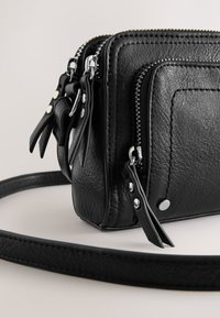 Črna usnjena torbica čez ramo z več žepki z zadrgo, teksturirano površino, srebrnimi okraski in nastavljivim trakom.