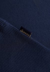 Tessuto a coste di colore blu navy con una finitura testurizzata, caratterizzato da un piccolo etichetta scura con lettering dorato che recita "Supersdry."