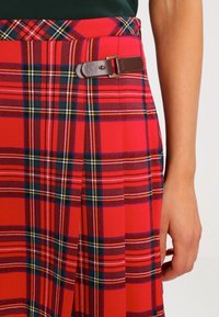 Jupe  à carreaux rouges présentant un mélange de motifs rouges vifs, bleu marine et jaunes. Comprend un détail de boucle en cuir marron à la taille.