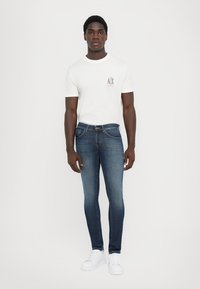 Vit bomull T-shirt med logotyp på bröstet, tillsammans med mörkblå skinny jeans och vita sneakers, bärs av en person som står mot en neutral bakgrund.