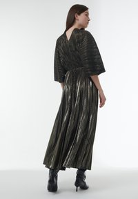 Robe noire longue plissée avec un dos en V profond et des manches larges, ornée d'accents en fil métallique. Portée avec des bottes à talons hauts noires.