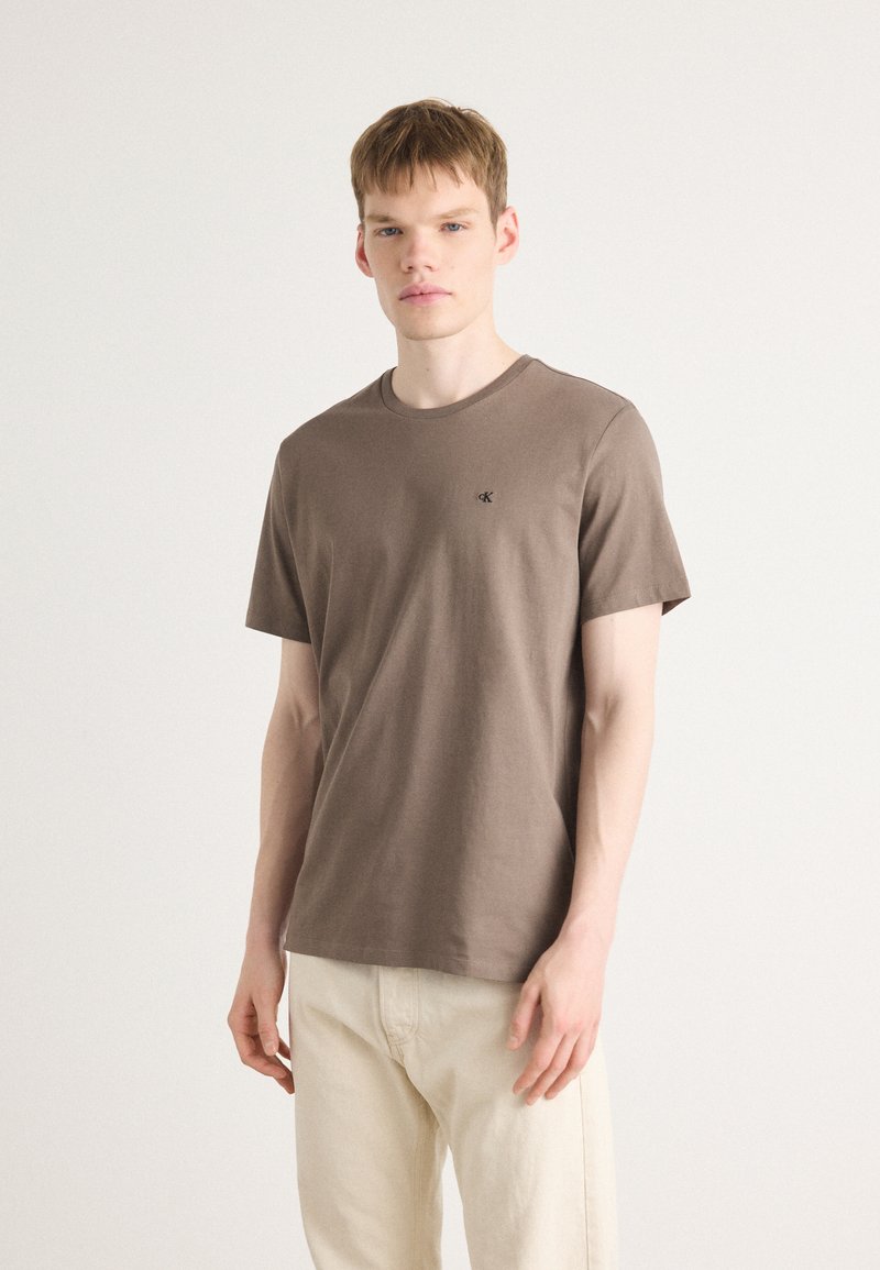 Braunes T-Shirt mit kurzen Ärmeln aus Baumwolle und Rundhalsausschnitt. Mit einem kleinen schwarzen Logo auf der linken Brust. Getragen mit hellen Hosen.