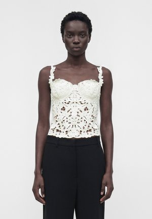 Czarna kobieta w białej koronkowej bluzce bez rękawów z kwiatowym wzorem i czarnych spodniach z wysokim stanem, stojąca na jednolitym jasnym tle.