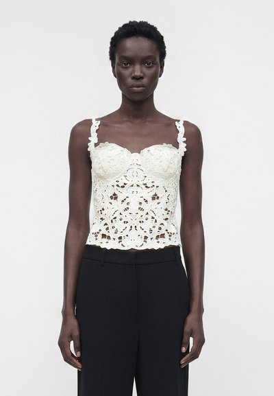 Femme noire portant un haut sans manches en dentelle blanche avec des motifs floraux et un pantalon noir taille haute, debout devant un fond clair uni.