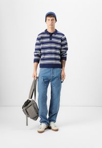 Marineblauwe en witte gestreepte trui met een kraag, blauwe spijkerbroek, witte sneakers en een grijze schoudertas. Model staat tegen een witte achtergrond.
