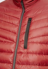 Giacca puffer rossa con cuciture orizzontali, colletto alto, zip frontale e una tasca con zip nera sul petto. Texture liscia e lucida.