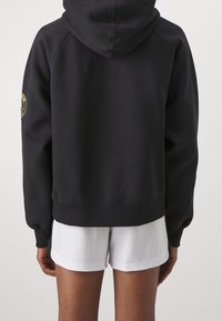 Sweat à capuche noir avec une grande capuche, des poignets et un ourlet inférieur côtelés ; présente un patch logo sur la manche gauche. Porté avec un short blanc.
