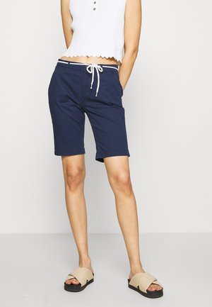 Mujer con camiseta blanca sin mangas y pantalones cortos azul marino hasta la rodilla con cordón blanco, de pie con sandalias beige de punta abierta sobre un fondo liso.