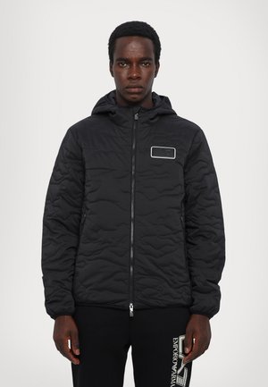 Veste matelassée noire avec capuche, dotée d'une fermeture éclair et d'un patch logo rectangulaire sur la poitrine. Poignets côtelés et texture lisse.