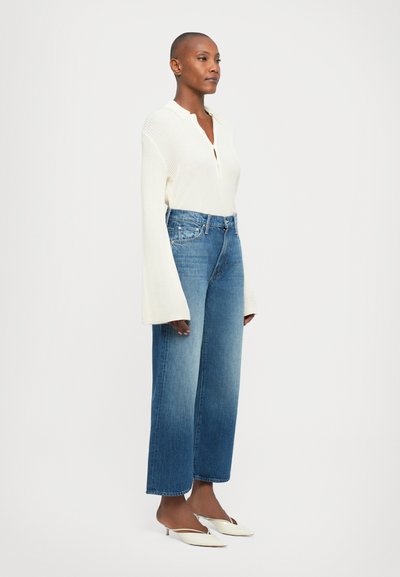 Vrouw met een geschoren hoofd die een crèmekleurige ribbeltrui met lange mouwen, hoge taille wijde blauwe jeans en witte puntige schoenen met lage hakken draagt.