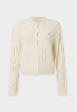 Femme aux longs cheveux bouclés portant un cardigan à carreaux crème et un pantalon large noir avec un motif en grille blanche, debout devant un fond uni.